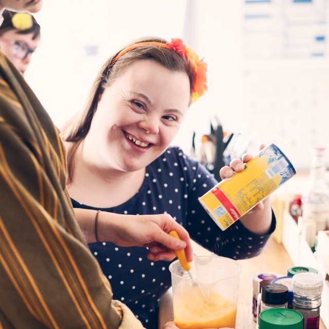 Eine junge Frau mit Downsyndrom und ihre Mitbewohner:innen kochen. Sie grinst fröhlich in die Kamera und salzt nebenbei das Essen