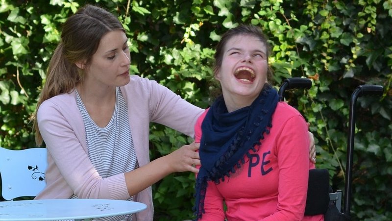 Zwei junge Frauen sitzen im Garten. Eine ohne Behinderung, eine mit schwerer Mehrfachbehinderung.
