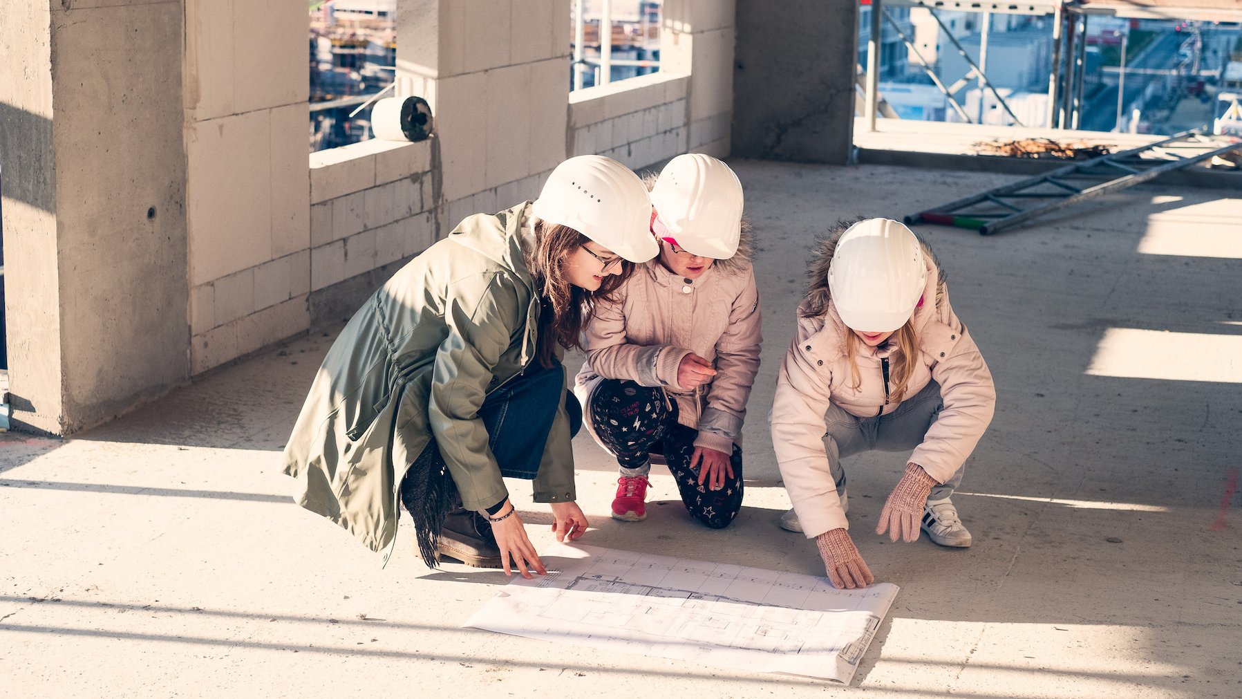 3 junge Frauen knien auf einem Bauplan. Sie befinden sich auf einer Baustelle und tragen weiße Bauhelme.