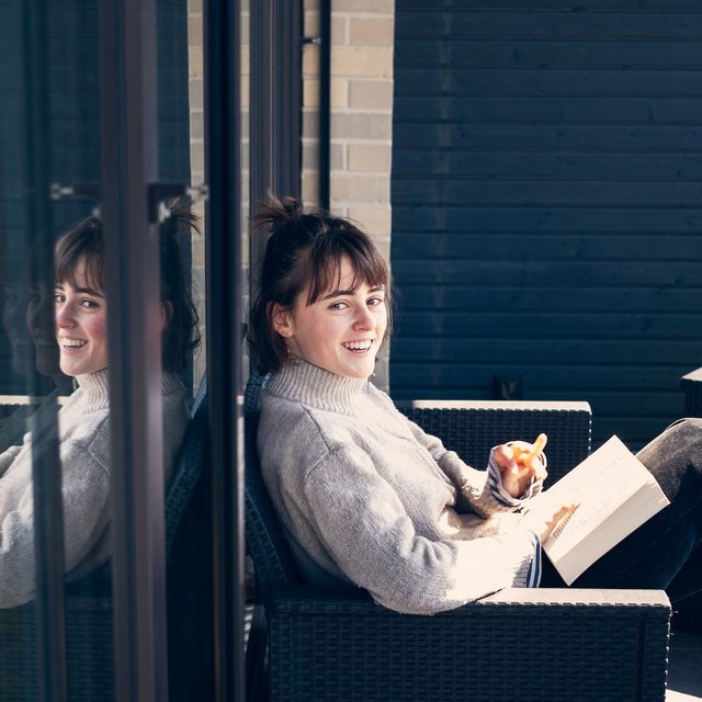Eine Mitbewohnerin einer inklusiven WG sitzt auf dem Balkon, lächelt in die Kamera und hält dabei ein Buch in der Hand