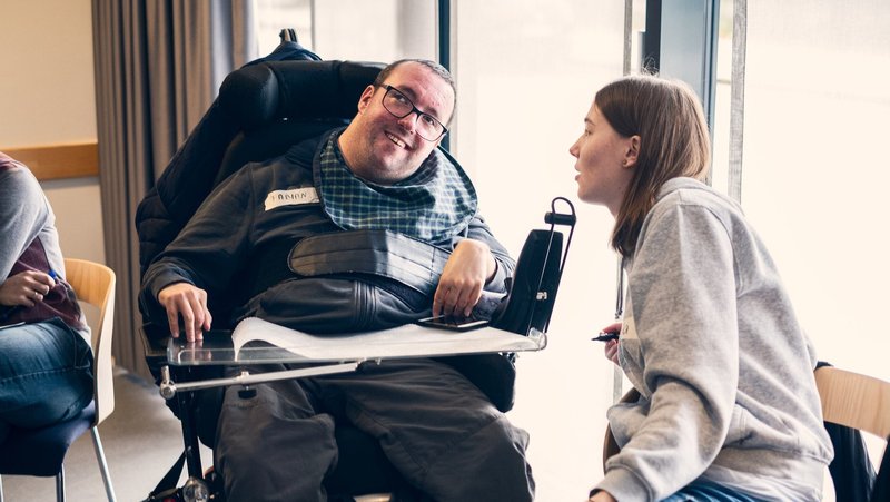 Ein junger Mann im Rollstuhl und eine junge Frau unterhalten sich. Sie lächeln 