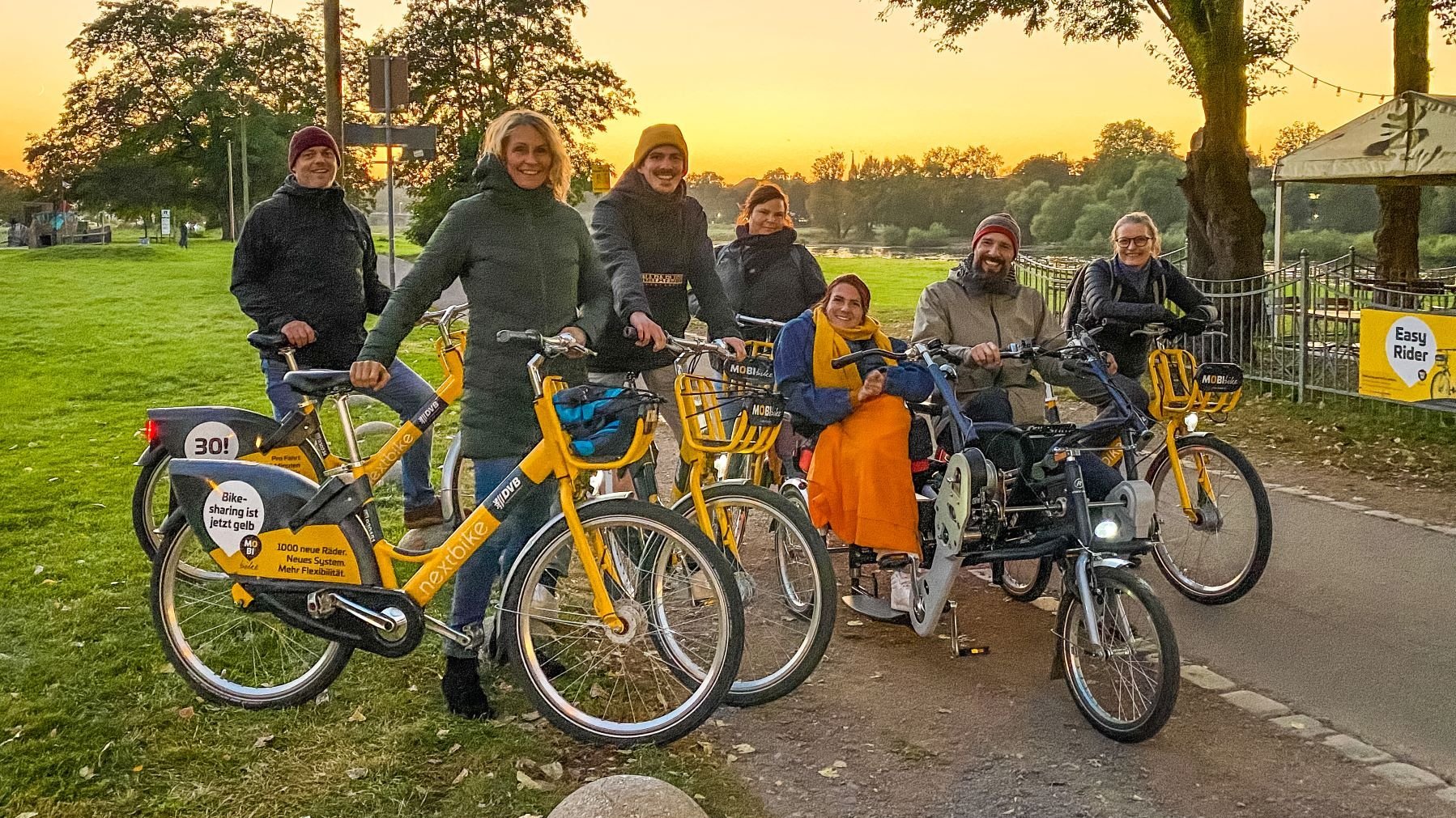Foto von einer Fahrradtour des WOHN:SINN-Teams. Alle stehen mit Fahrrädern vor einem Sonnenuntergang. Zwei Personen sitzen in einem Rikscha.