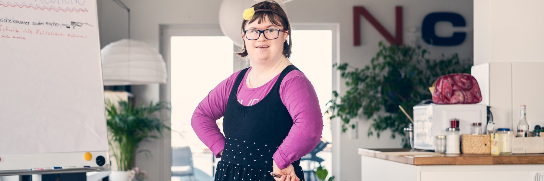 Eine junge Frau mit Downsyndrom steht in der WG-Küche neben einem Flipchart und blickt entschlossen in die Kamera